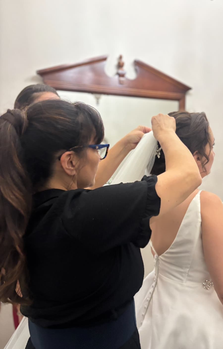The image shows a hairstylist standing behind a bride, adjusting and securing a white veil into an updo hairstyle. The bride is wearing a white sleeveless wedding dress with a buttoned back and subtle decorative detailing, facing away from the camera. The stylist’s hands are raised, actively positioning the veil, indicating a candid, in-progress preparation moment rather than a posed shot. The indoor setting is softly lit with neutral walls and a wooden-framed mirror in the background, suggesting a bridal suite or dressing room environment suitable for behind-the-scenes or service-in-action website placement.