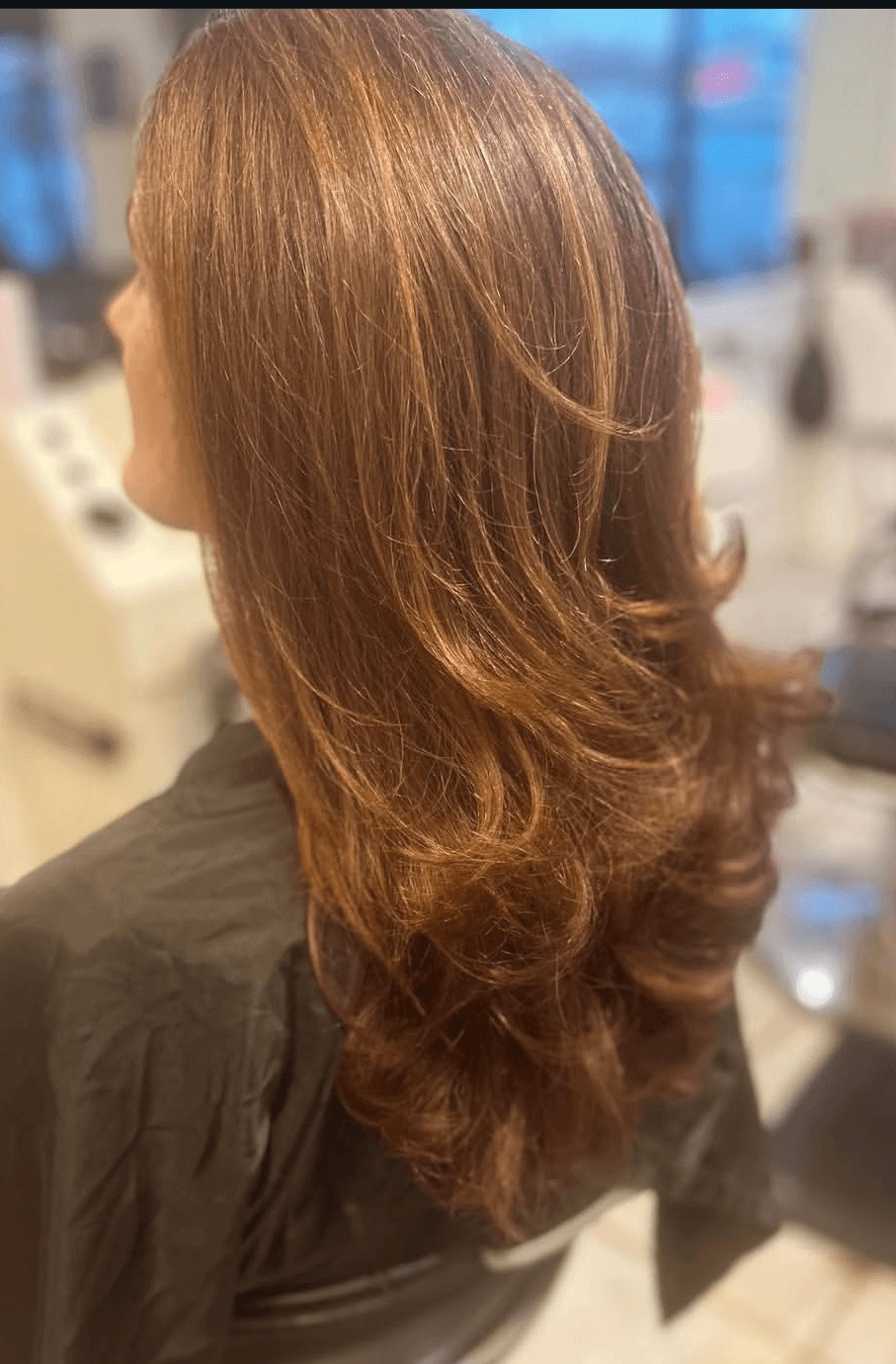 The image shows a close-up, angled side view of a finished hairstyle on a client seated in a salon chair. The hair is medium-to-long length, warm auburn or copper-toned, styled with smooth layers and soft, outward-curving ends that add volume and movement. Lighting highlights the natural sheen and color variation throughout the hair, while the background is softly blurred, showing indistinct salon tools and stations. The composition focuses on texture, color, and shape, making it suitable for a portfolio, service results, or before-and-after showcase on a hairstylist’s website.