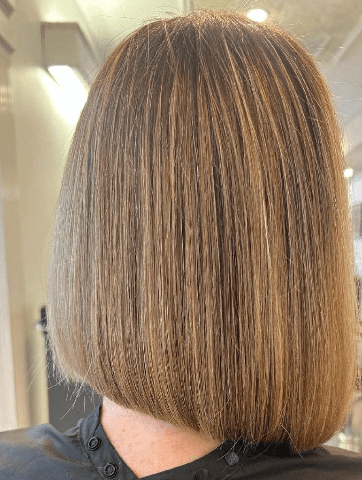 The image shows a rear view of a completed blunt bob haircut on a client seated in a salon chair. The hair is cut to just above shoulder length with a clean, straight perimeter and precise edge alignment, creating a sharp, structured silhouette. Subtle, evenly distributed highlights add dimension and depth to the medium brown base color, visible under bright salon lighting. The background is softly out of focus with salon fixtures and mirrors, keeping attention on the cut’s shape, finish, and color accuracy, making it suitable for a haircut portfolio or style results gallery.